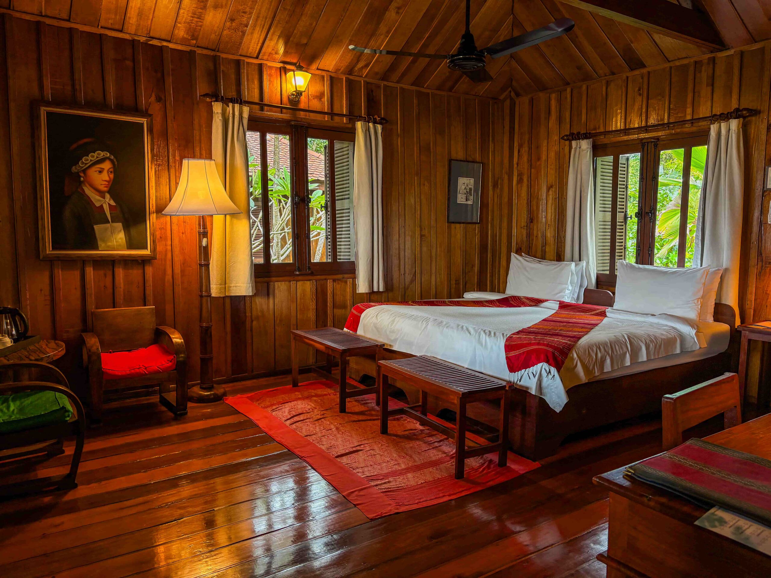 Bungalow bedroom with double beds, a high timber ceiling, red woven textiles and a framed portrait beside a floor lamp